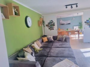 Bild vom Wohnzimmer der Wohngruppe. Es zeigt eine grüne Wand mit einem grauen Sofa an der Wand stehend. Auf dem Sofa liegen mehrere bunte Kissen. Im Hintergrund ist der Esstisch erkennbar.